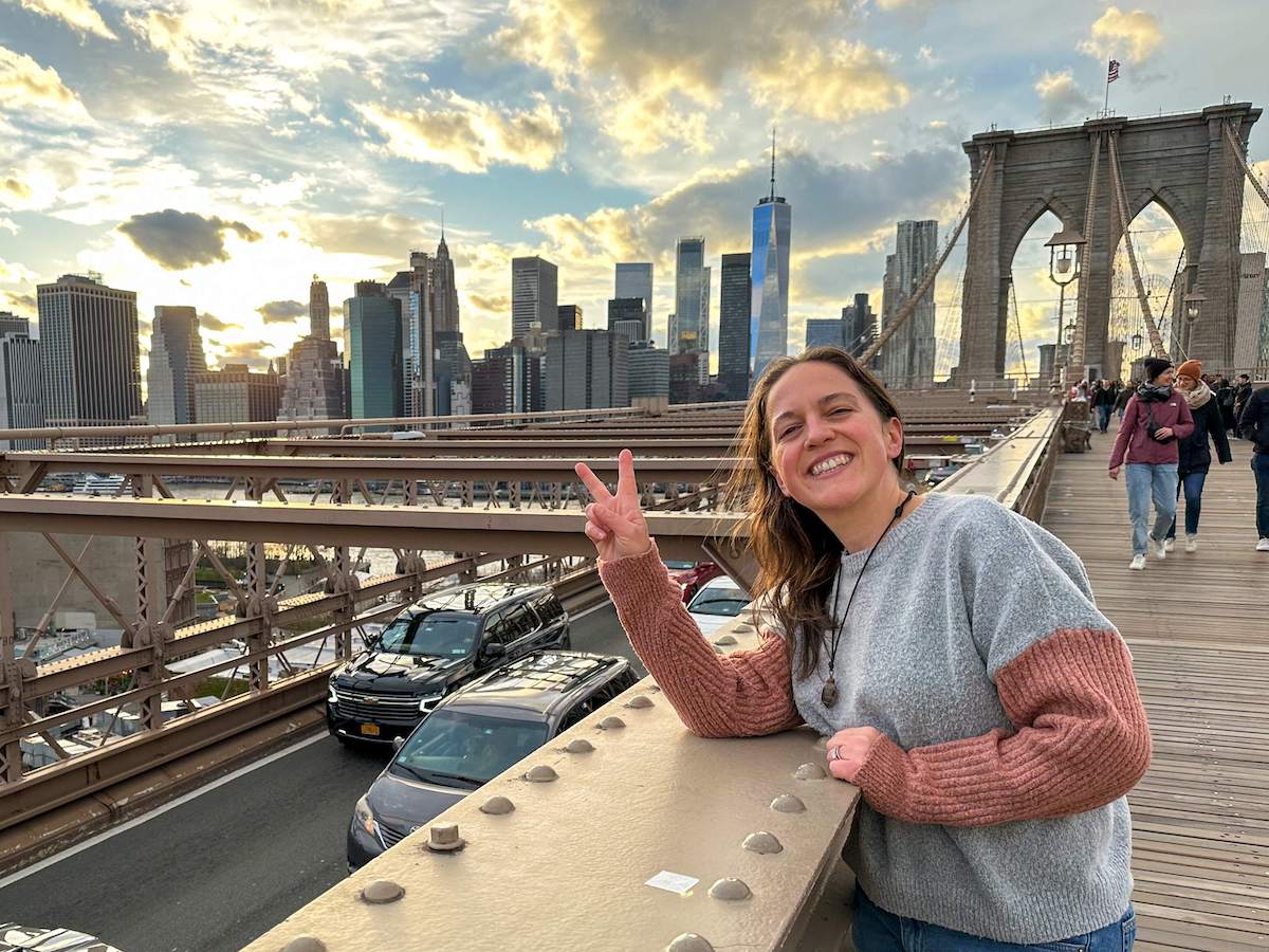 "A woman walking on the Brooklyn Bridge from DUMBO, Brooklyn to Lower Manhattan at sunset, with the city skyline and the bridge's structure visible in the background.