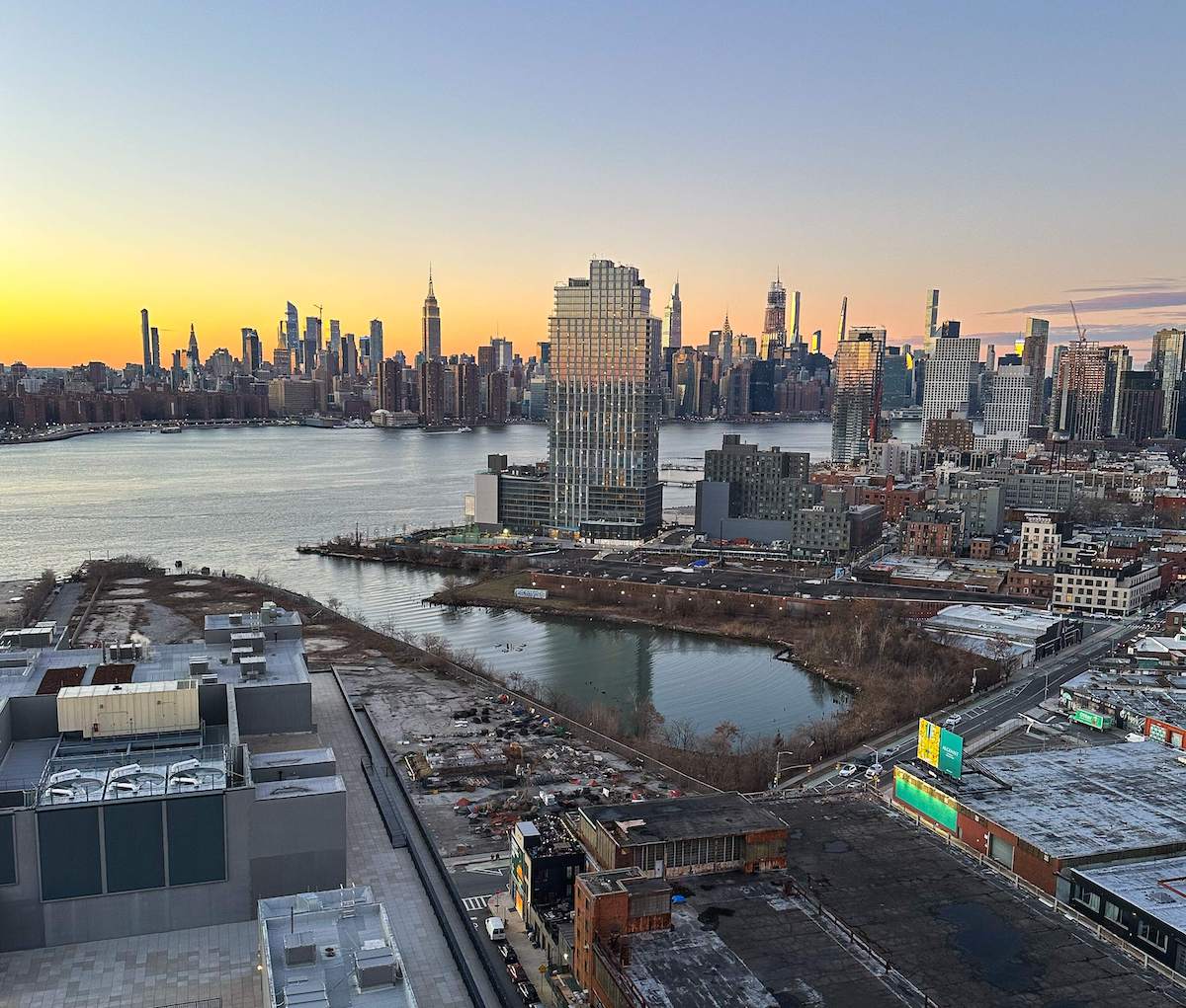 A view of the Manhattan skyline at sunset from the Westlight rooftop bar in Brooklyn, overlooking the East River and various buildings, with a soft orange and blue sky in the background.