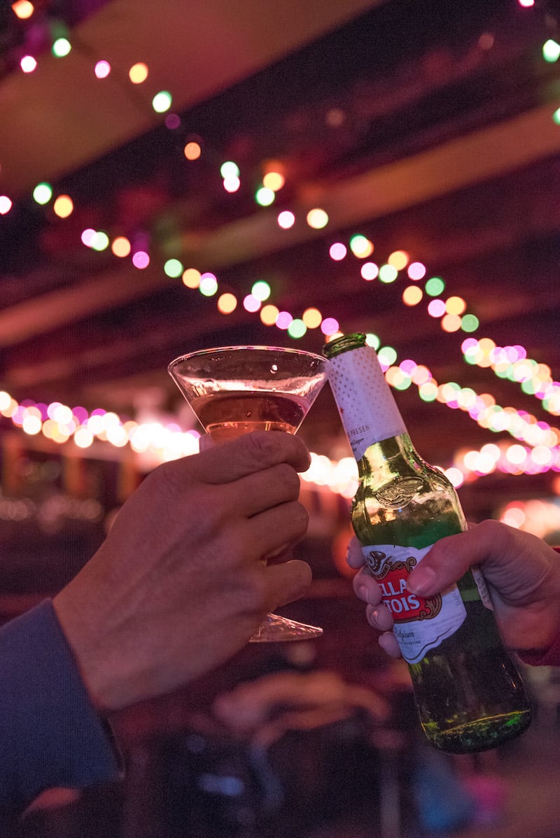 Two people clinking glasses at Marie's Crisis bar during a night out in New York City, with colorful string lights creating a festive atmosphere in the background.