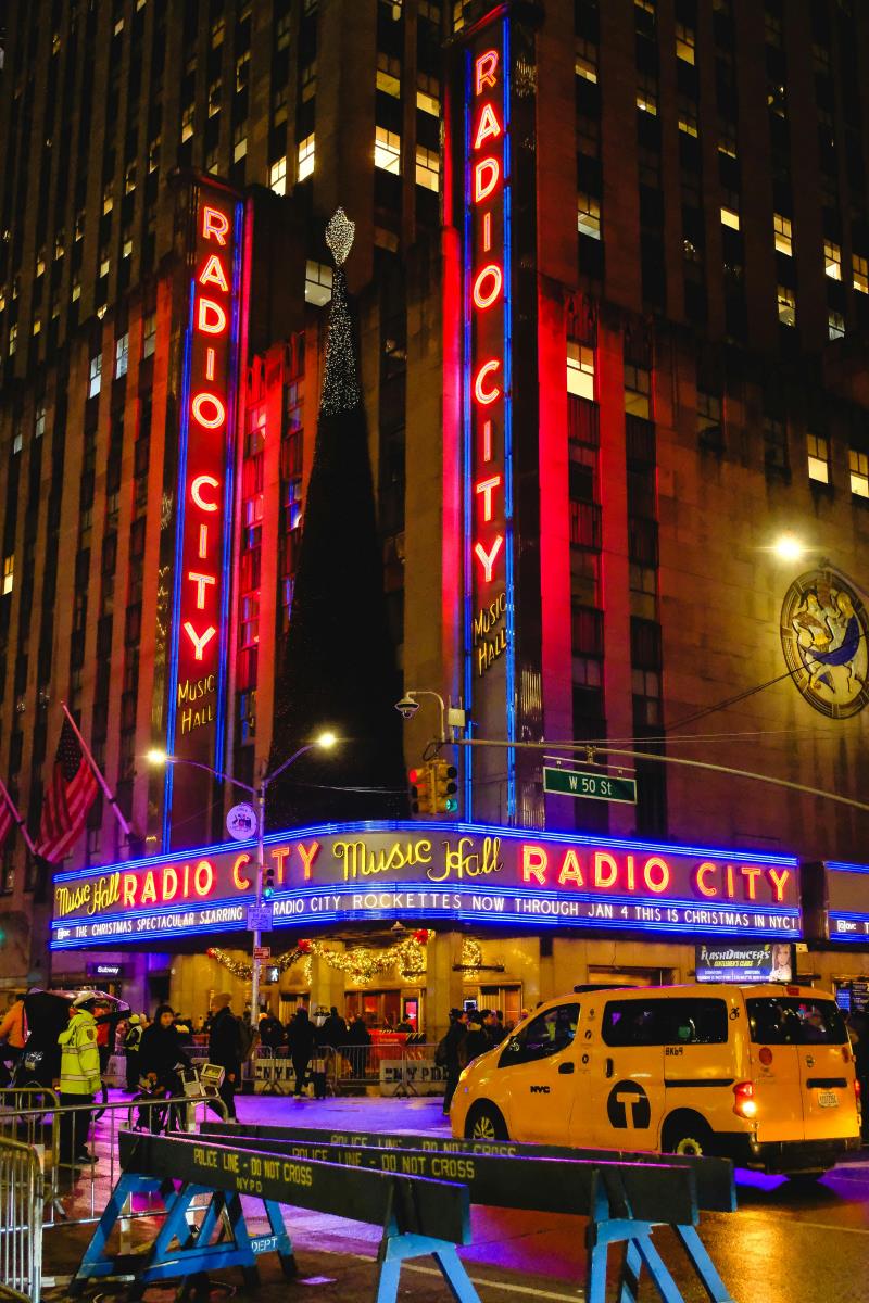 Radio City Music Hall in NYC at night, brightly illuminated with neon lights and decorated for the holiday season, with people and a yellow taxi in the foreground.