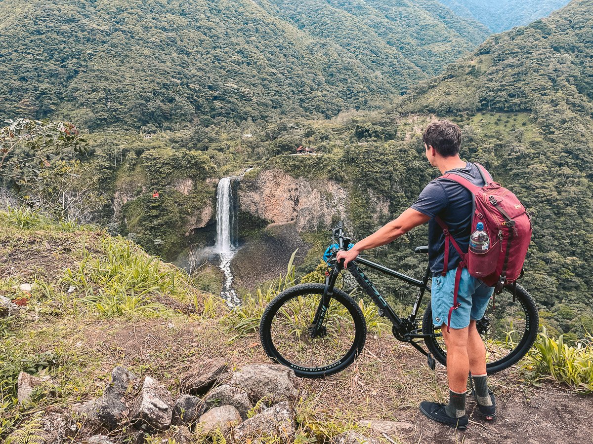 A solo traveler with a bike overlooking the Cascada Manto de la Novia waterfall during the Ruta de las Cascadas in Baños, Ecuador, surrounded by verdant mountains, highlighting the adventurous and scenic aspects of solo travel in Ecuador.