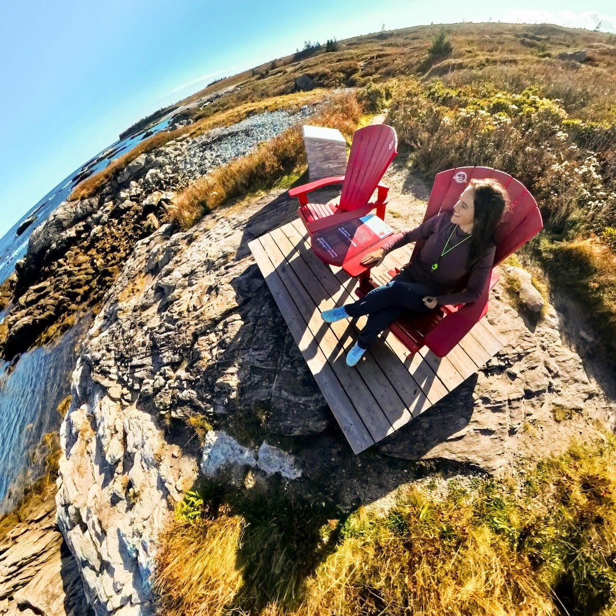 Woman relaxing in a red Adirondack chair at Kejimkujik National Park Seaside, overlooking rocky shores and grassy coastal landscapes, with bright sunshine casting shadows on the wooden platform.