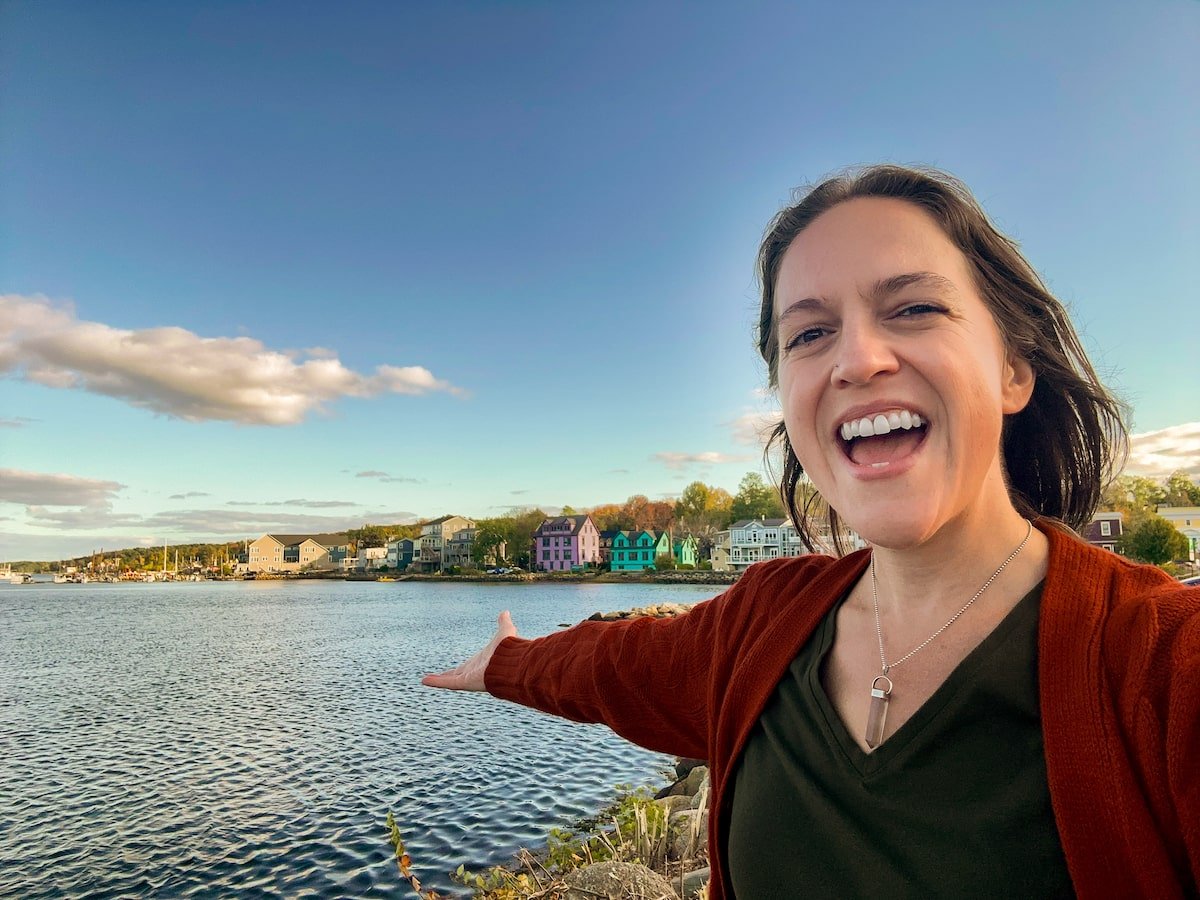 Smiling woman taking a selfie with the colorful houses along the Mahone Bay waterfront in the background, with calm water and a clear sky completing the picturesque scene.