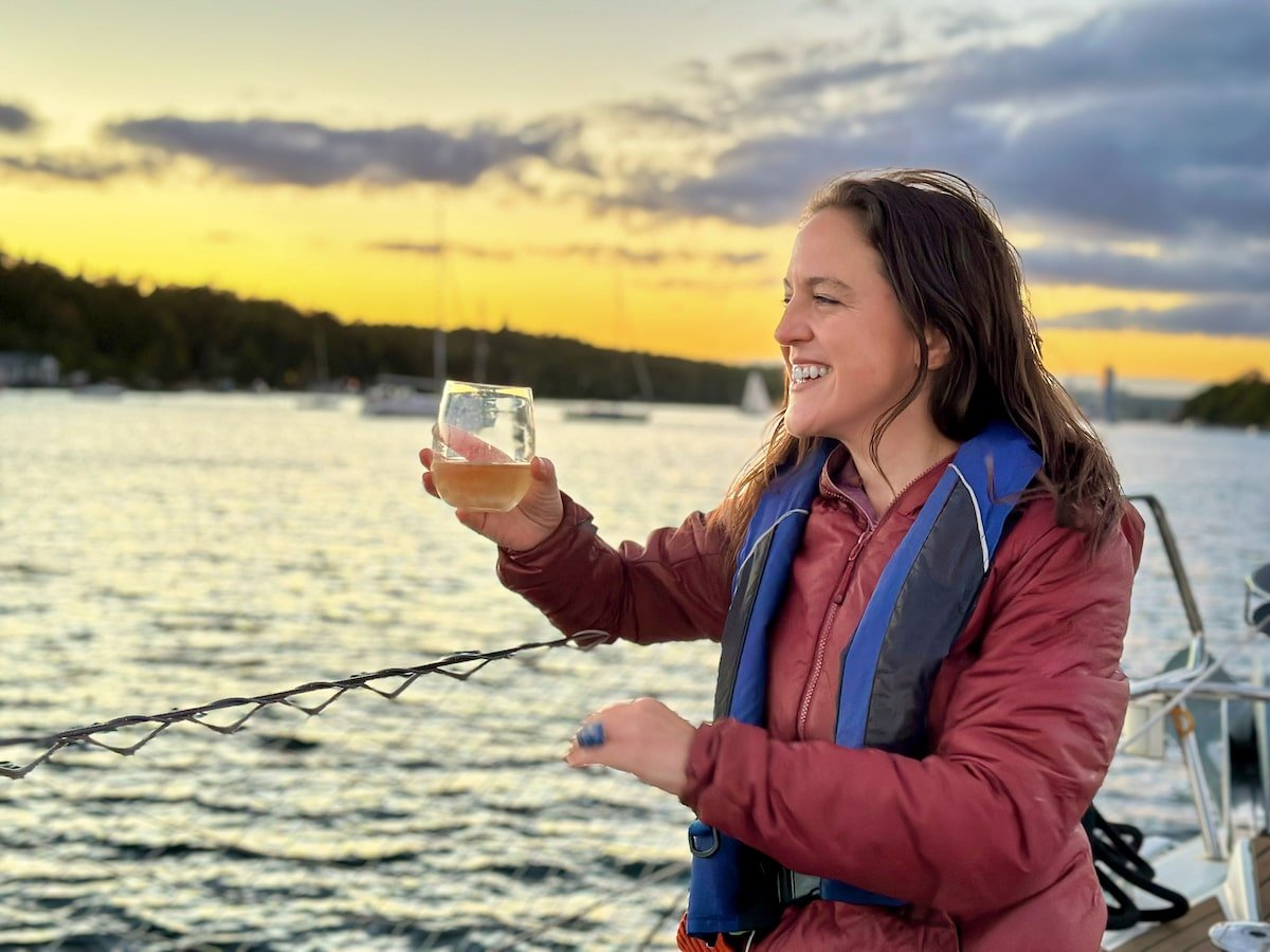 Person smiling and holding a glass of wine while enjoying a Sunset Wine & Cheese Sail with J Farwell Sailing Company in Halifax, Nova Scotia, with the sun setting over the water in the background.