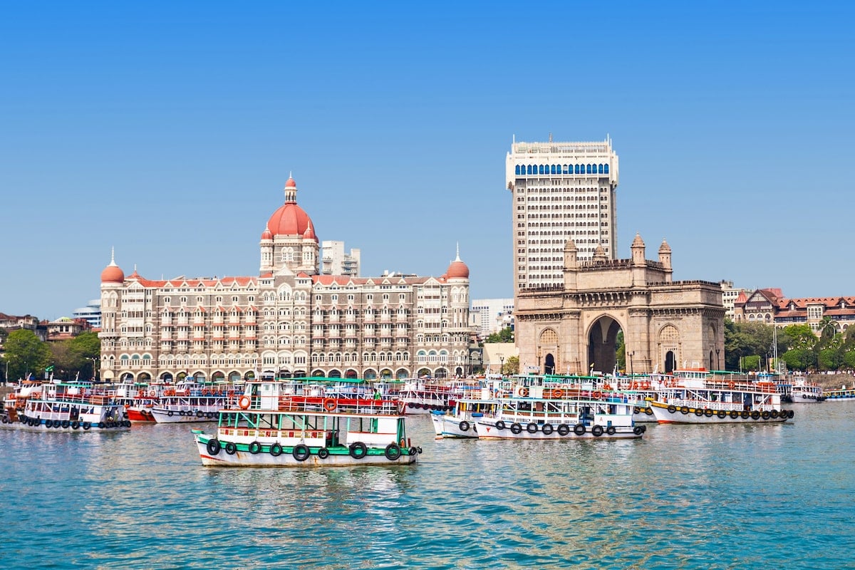 View of the Taj Mahal Palace Hotel and Gateway of India in Mumbai, with vibrant ferry boats docked in the sparkling blue waters of the Arabian Sea under a clear blue sky.