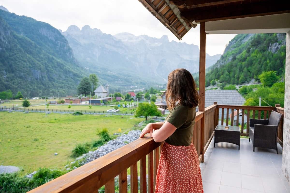 Woman standing on a wooden balcony at Bujtina Polia guesthouse in Theth, Albania, overlooking green pastures and dramatic mountain peaks in the distance.