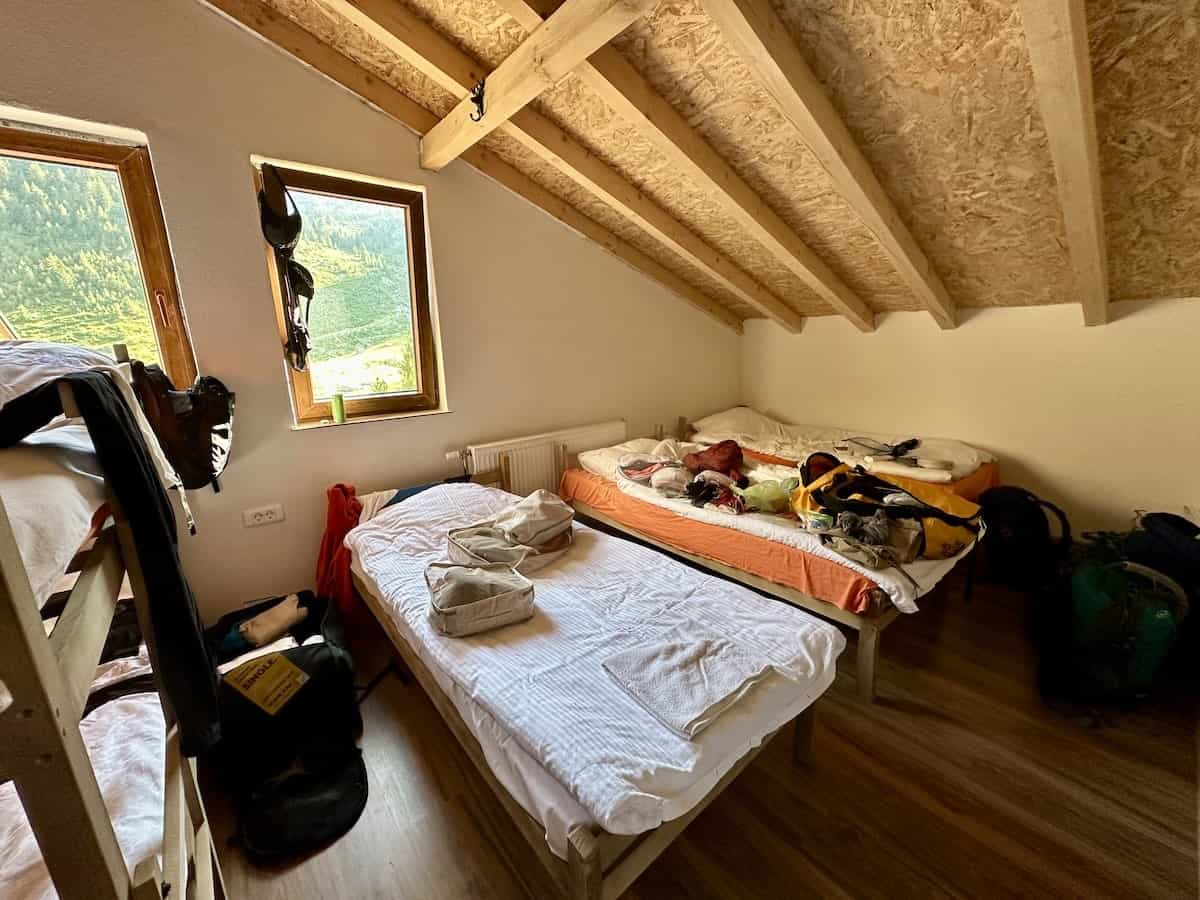 A simple guesthouse room with wooden ceiling beams, bunk beds, and two single beds, one with hiking gear spread across it, at Gacaferi Guesthouse in Deçan, Kosovo.