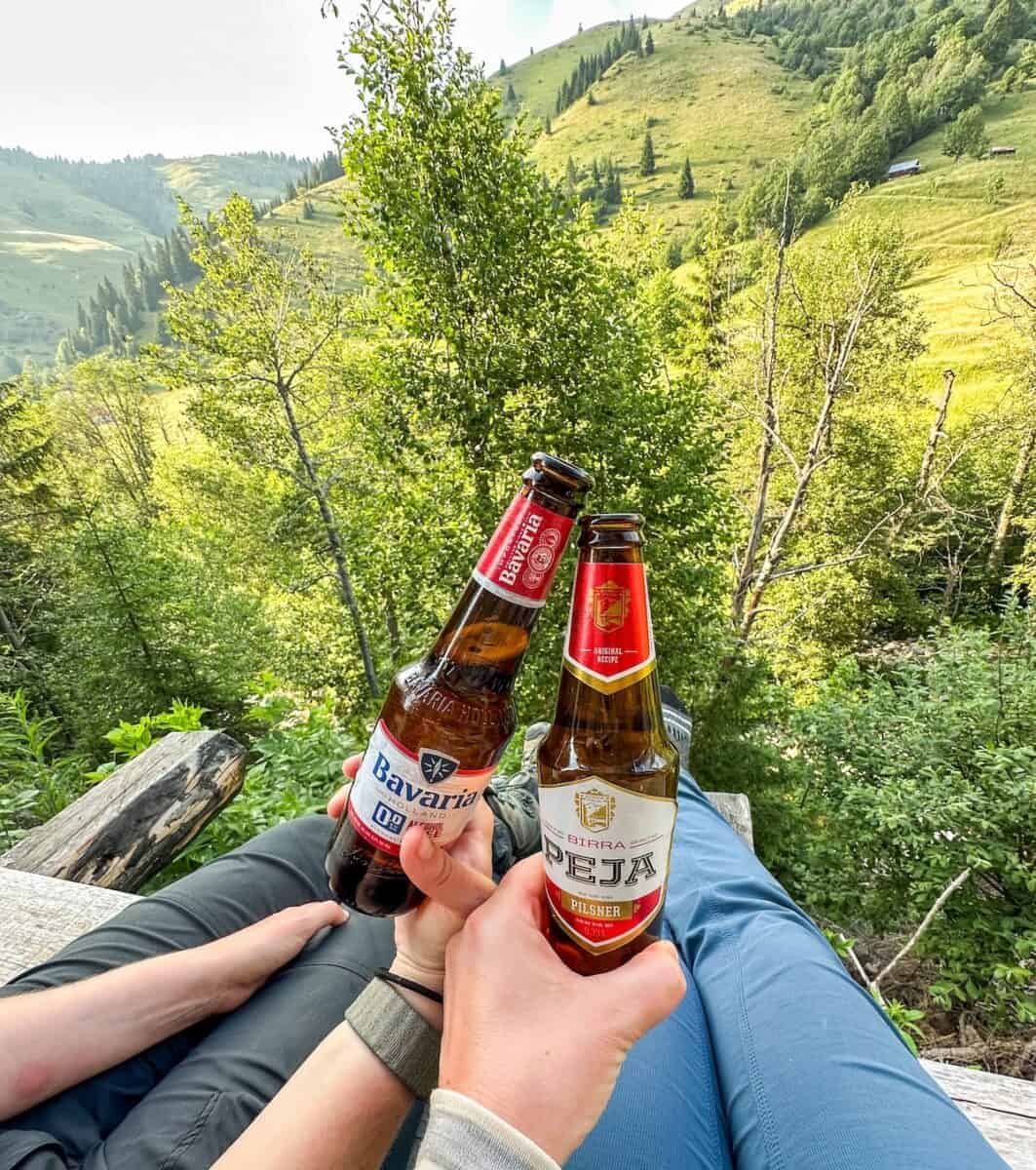 Two hikers toast with bottles of beer while relaxing at “HRID” Eco Mountaineering Village in Babino Polje, Montenegro, with lush green hills and trees stretching into the distance.
