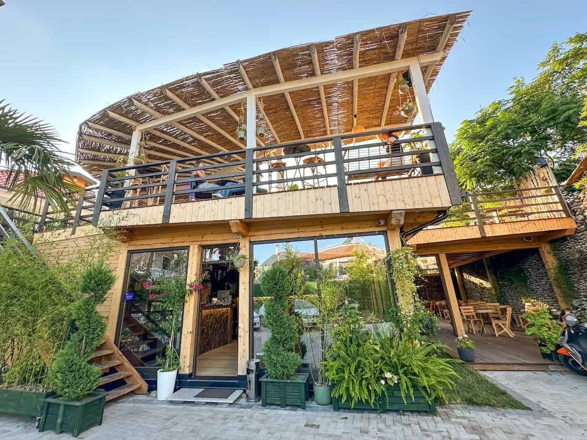 The stylish wooden exterior of Hotel Ikona in Shkodër, Albania, featuring its restaurant with lush greenery and a rooftop dining area under a woven canopy.