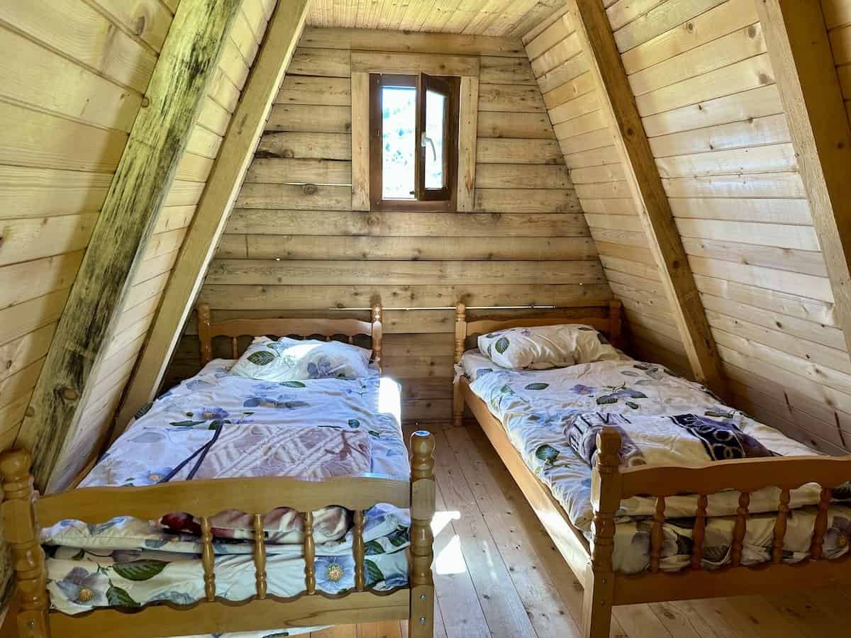 A cozy wooden A-frame guesthouse room with two single beds, floral bedding, and a small window letting in natural light at Leonardi Guesthouse in Doberdol, Albania. Ask ChatGPT