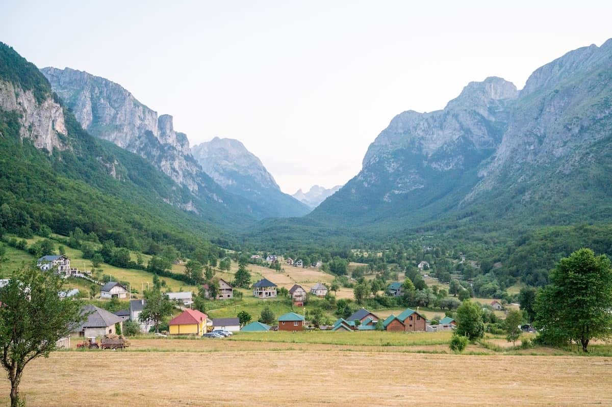 A peaceful view from Moonlight House in Vusanje, Montenegro, showing a village with colorful rooftops nestled in a wide green valley, surrounded by dramatic mountain peaks under a pale sky. Ask ChatGPT