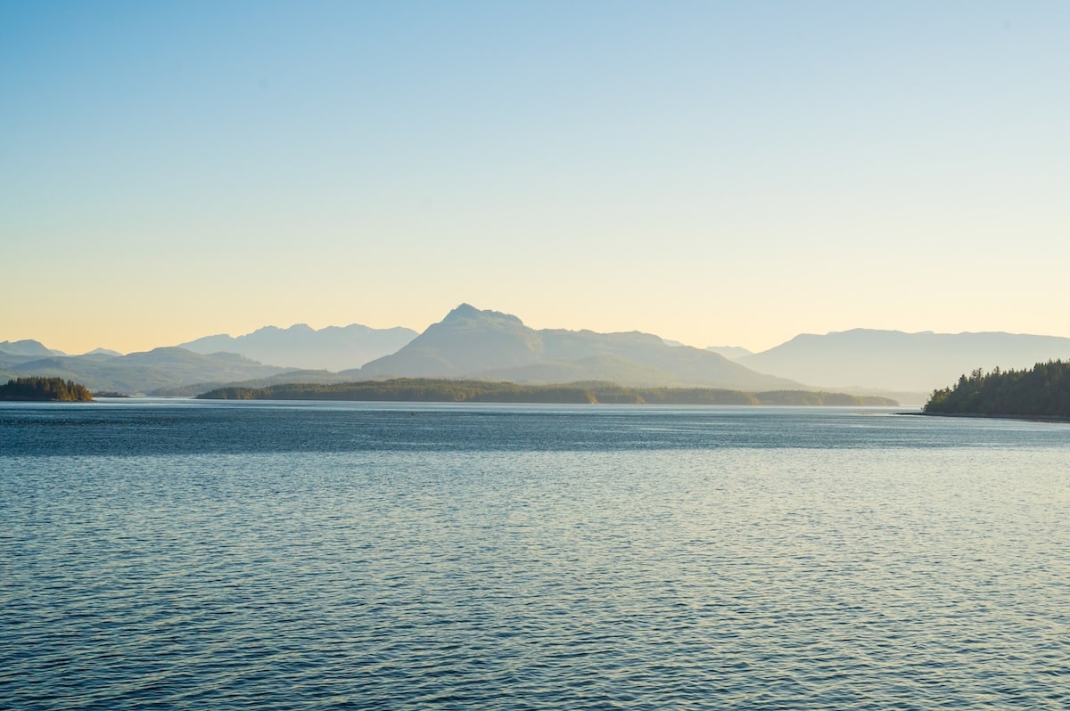 Golden hour view of Alaska’s Inside Passage with layered mountain silhouettes and calm blue water, photographed from the bow of a cruise ship.