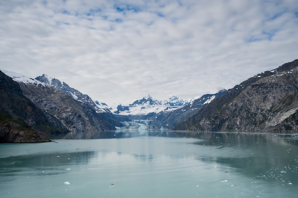 Scenic view of Johns Hopkins Glacier and surrounding snow-capped peaks from Jaw Point in Glacier Bay National Park, Alaska, with icy turquoise waters and dramatic rocky cliffs under a partly cloudy sky.