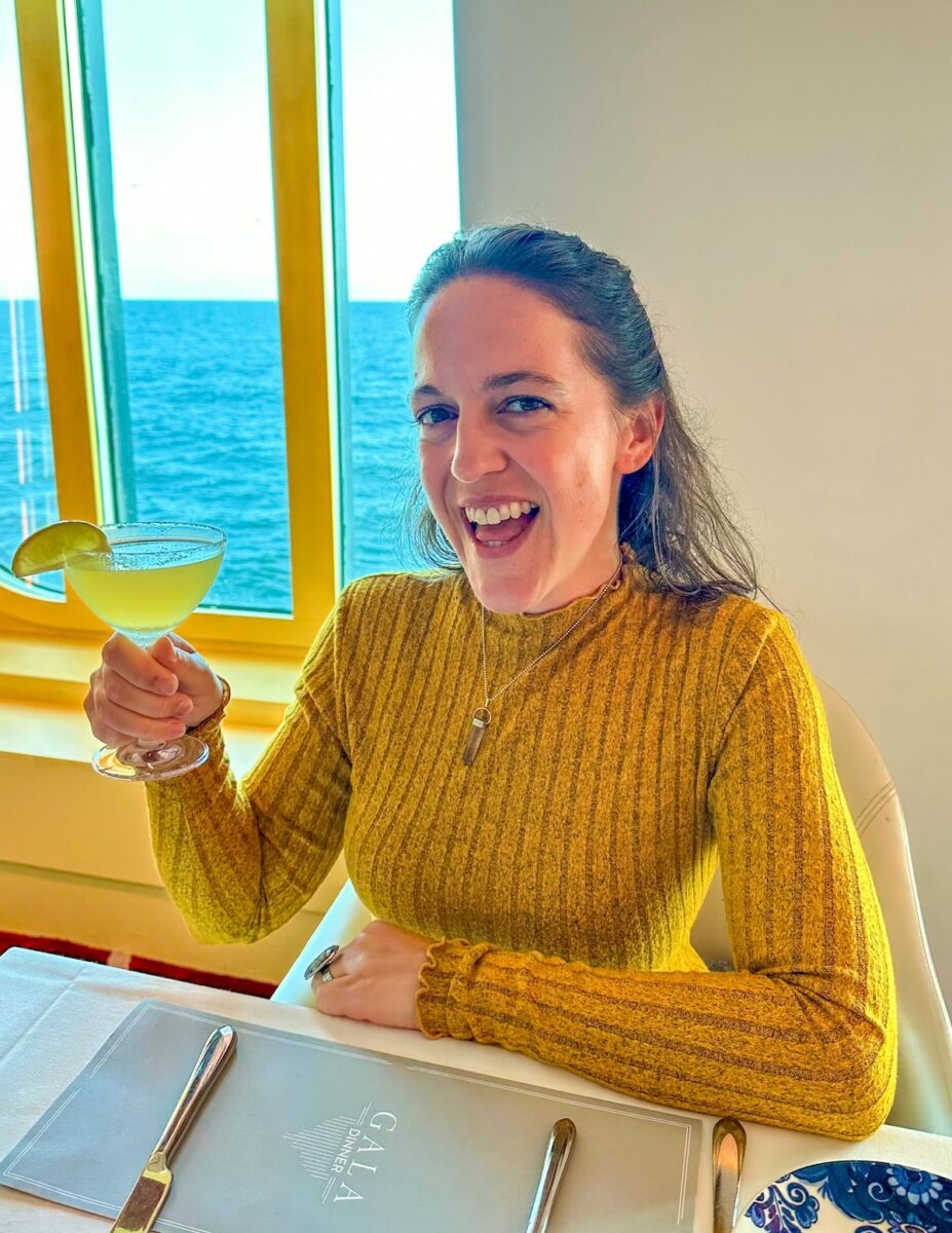 Woman enjoying a cocktail during Gala Night in the Main Dining Room on Holland America’s Koningsdam Alaska cruise, seated by a window with ocean views in the background.