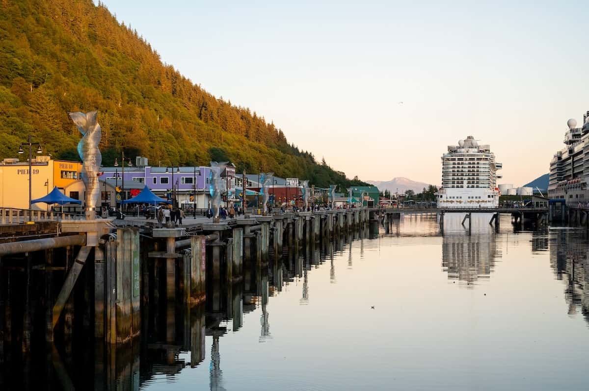 Cruise ships docked at the Juneau, Alaska port with colorful waterfront buildings, mountains rising behind, and calm water reflecting the scene at sunset.
