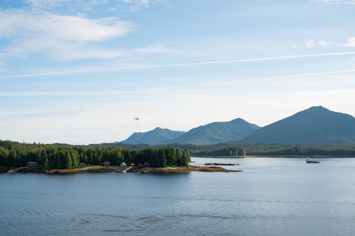 Scenic view of forested islands and distant mountains as the cruise ship sails away from Ketchikan, Alaska, with a small seaplane flying above the calm blue water.