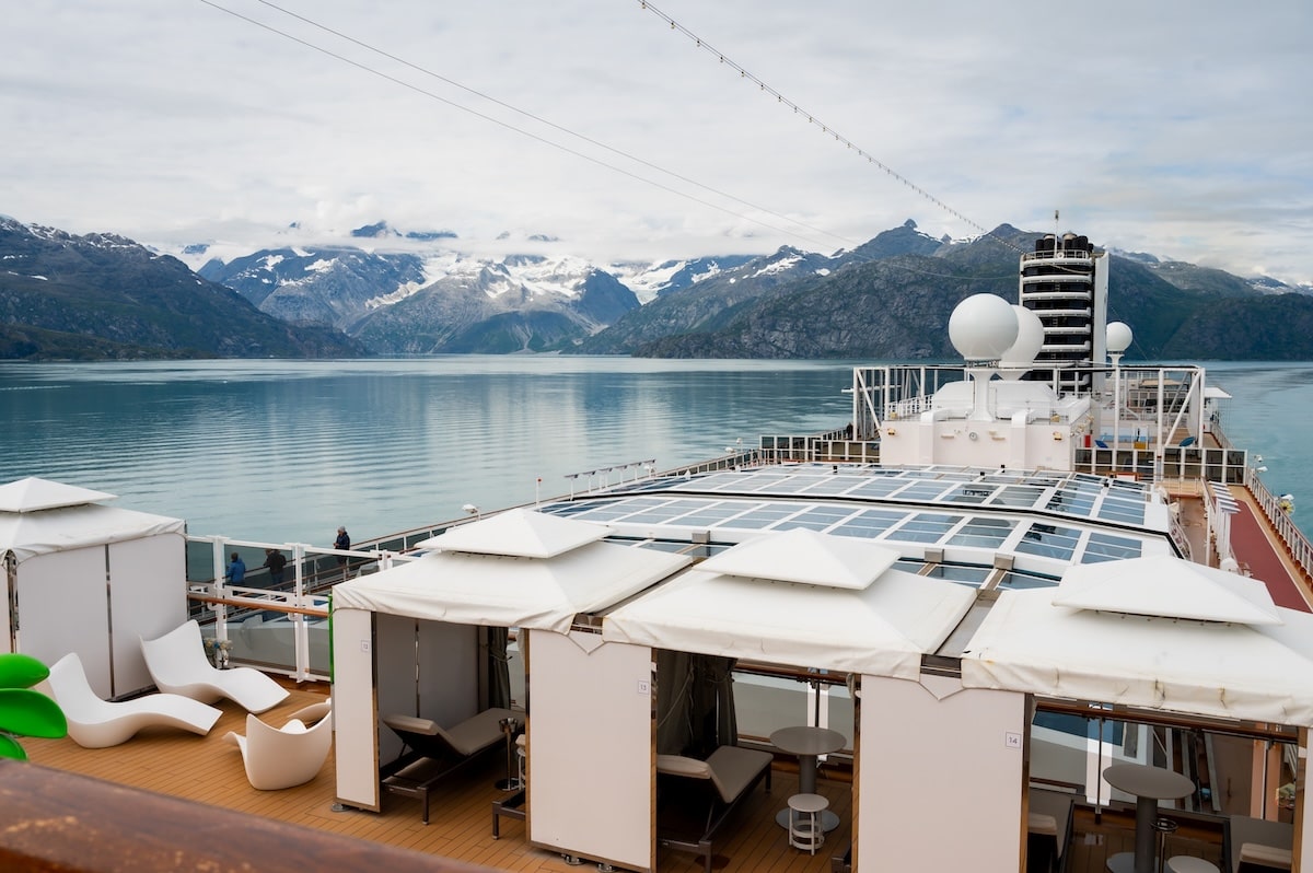 View of Glacier Bay’s snow-capped mountains and calm turquoise water from the upper deck of Holland America’s Koningsdam cruise ship, with private cabanas and lounge chairs in the foreground.