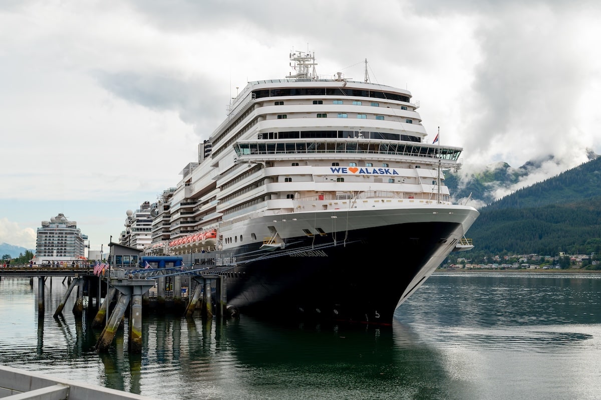 Front view of the Holland America Koningsdam cruise ship docked at the port in Juneau, Alaska, with a ‘We ♥ Alaska’ banner displayed across the upper deck and misty forested mountains in the background.
