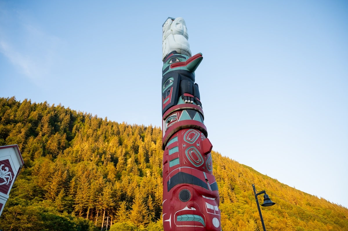 A vividly painted Haida Eagle totem pole (Gyáa'aang Kootéeyaa Deiyí) carved by artist Warren Peele stands tall against a backdrop of forested hills at sunset along Juneau’s Totem Pole Trail in Alaska.