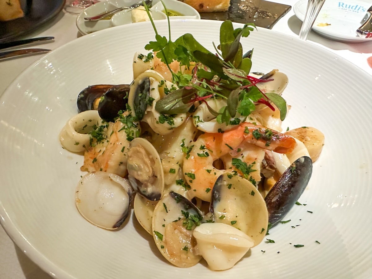 Seafood pasta dish at Rudi’s Sel de Mer on Holland America’s Koningsdam, featuring shrimp, clams, and mussels tossed with herbs and garnished with fresh greens in a white bowl.