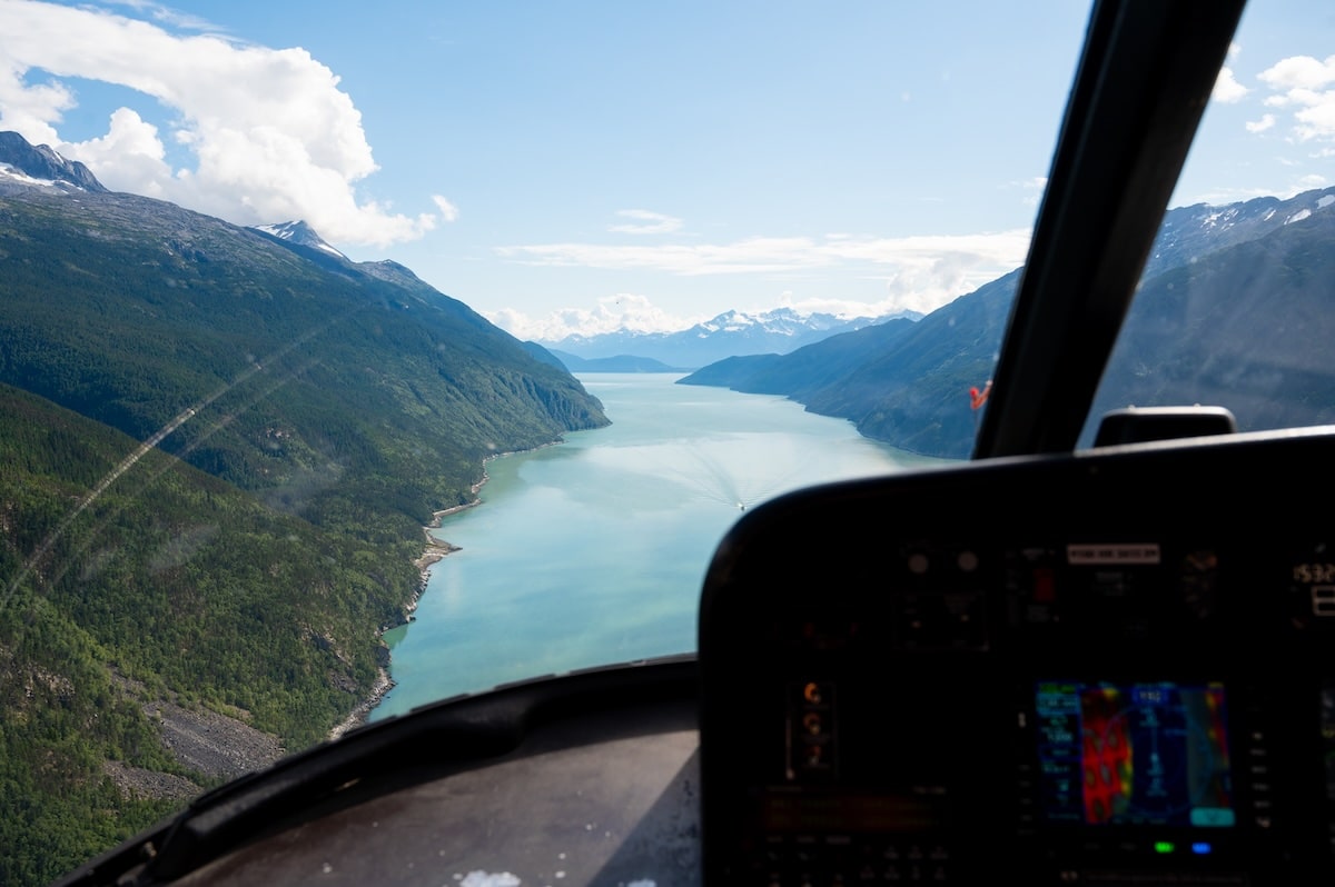 Scenic aerial view from inside a helicopter flying over the turquoise waters and forested mountains of Skagway, Alaska, with the helicopter dashboard visible in the foreground and distant snow-capped peaks under a bright blue sky.