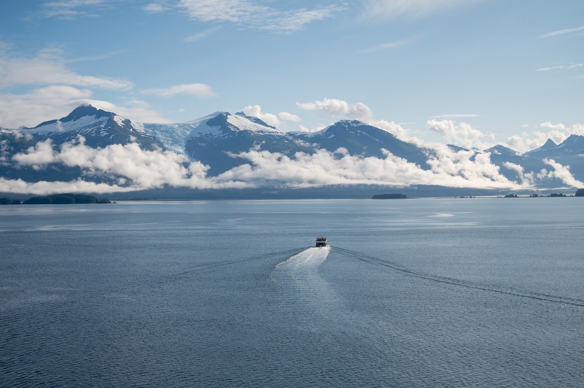 A small excursion boat sails away from the Holland America Koningsdam through Alaska’s Tracy Arm Fjord, surrounded by deep blue water, misty clouds, and snow-capped mountains.