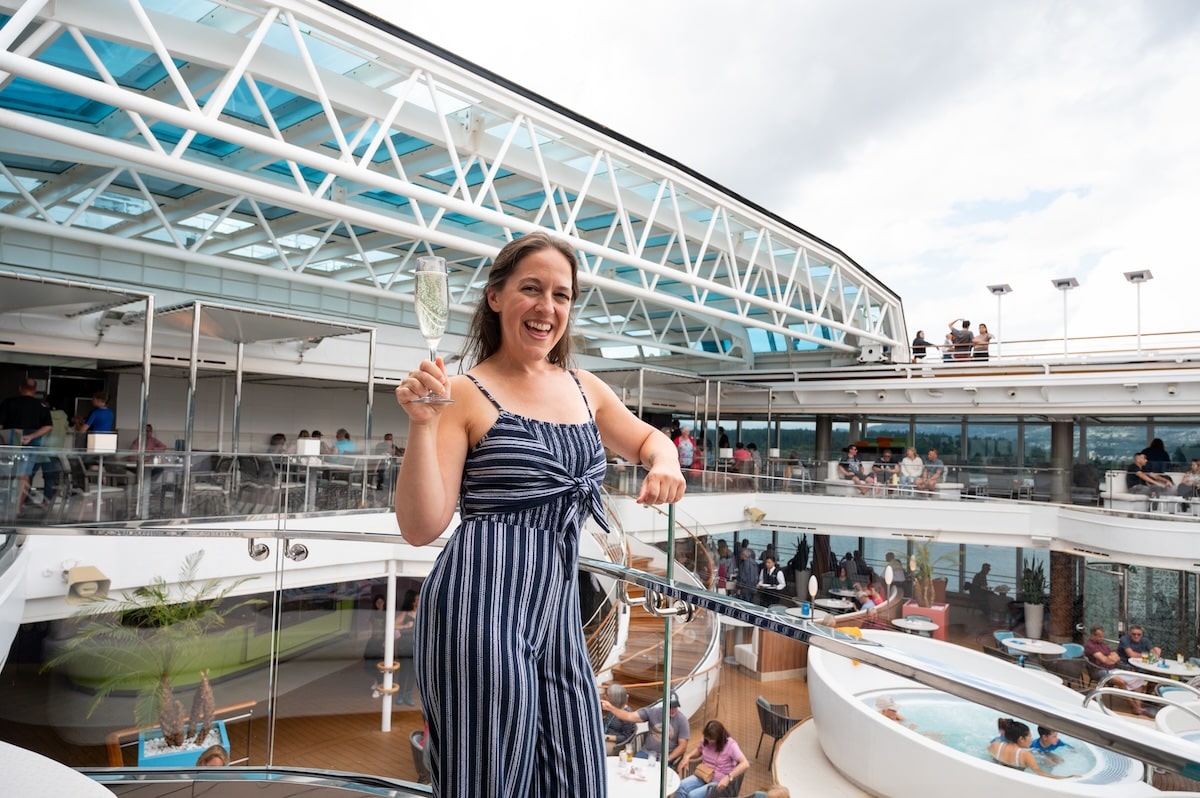 Woman in a navy and white striped jumpsuit smiling and holding a glass of champagne on the Holland America Koningsdam’s Lido Pool deck, with people relaxing in the background.